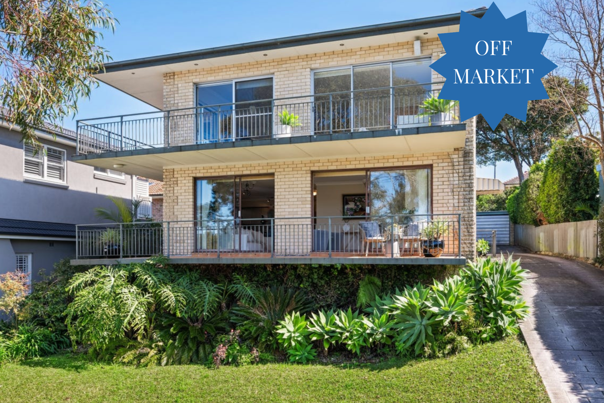 Two-story brick house with a spacious balcony and metal railing, surrounded by green shrubs and palm-like plants; blue OFF MARKET badge on the right side.