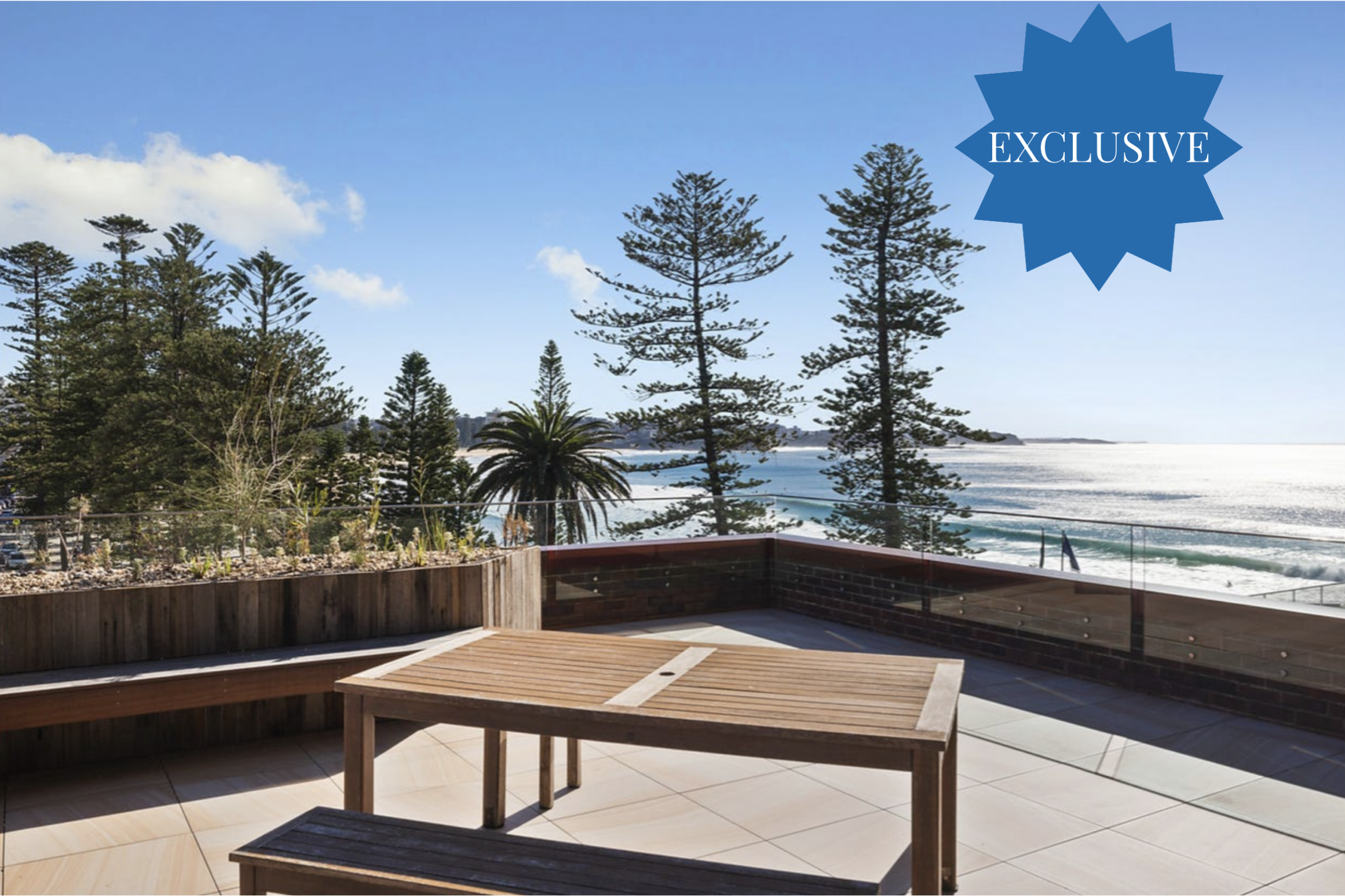 Patio scene with a wooden table and benches overlooking the ocean, with tall trees and a clear blue sky; a blue 'Exclusive' badge overlays the upper-right corner.