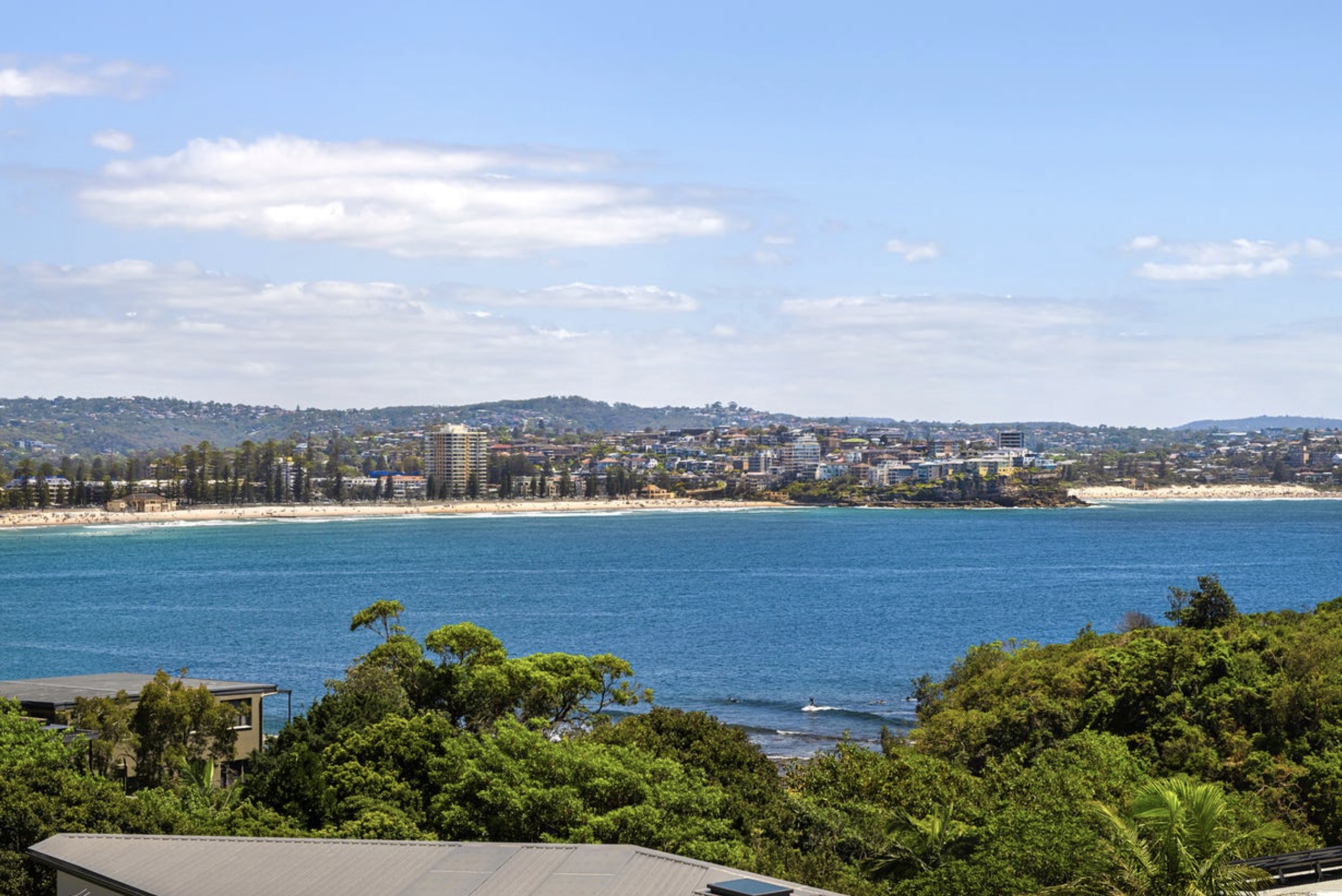 Buyers Agent seaforth– coastal bay view with green trees, sandy beach, town buildings and transport under a cloudy sky.