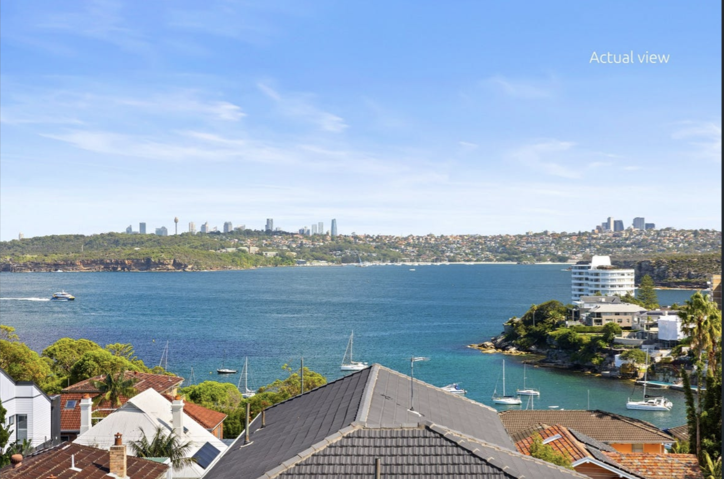 Coastal town by the bay with boats, residential rooftops, and a distant city skyline under a blue sky.
