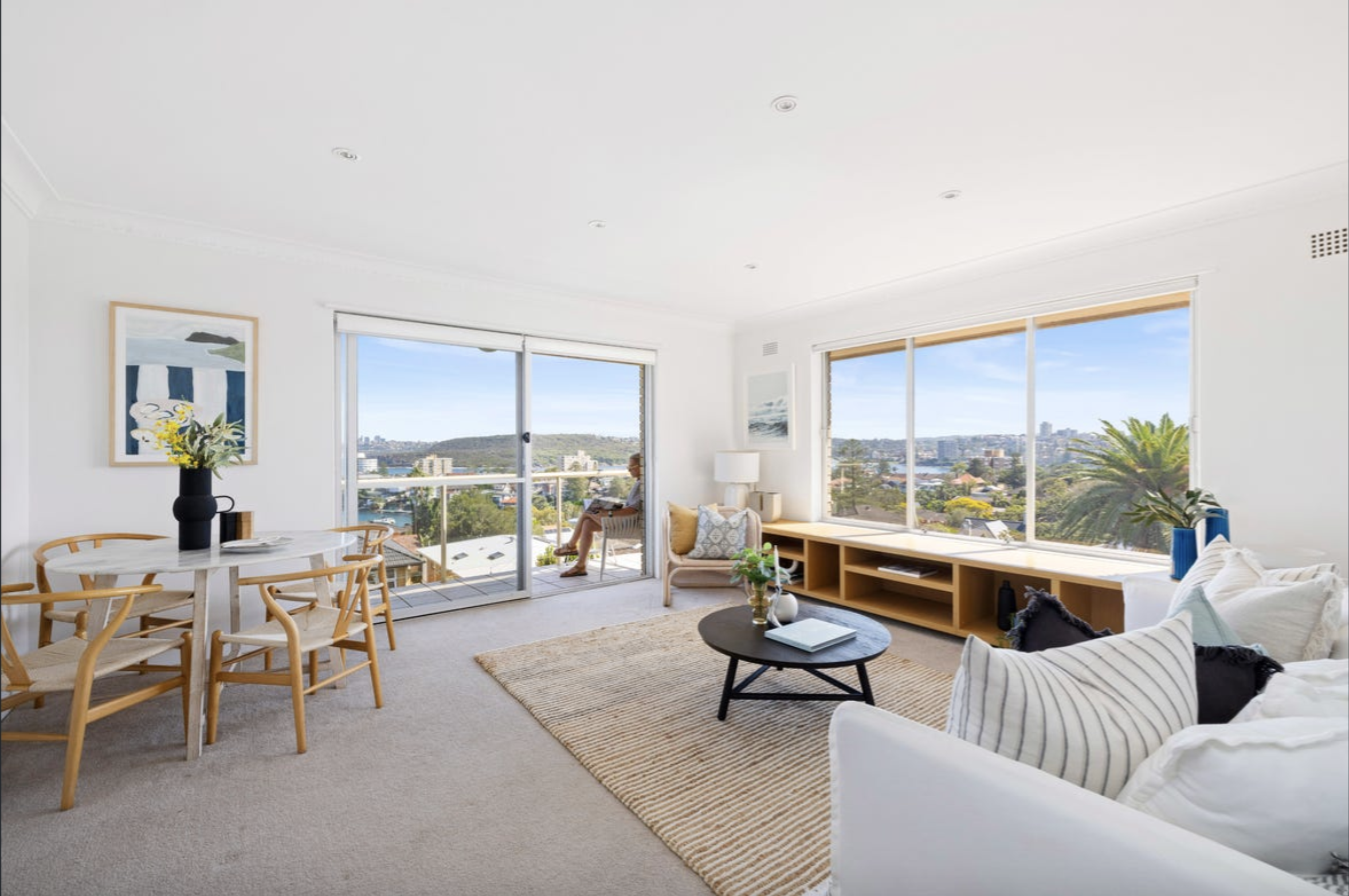 Bright, modern living room with large windows offering a city and water view; light furniture and a beige rug over carpeted floor