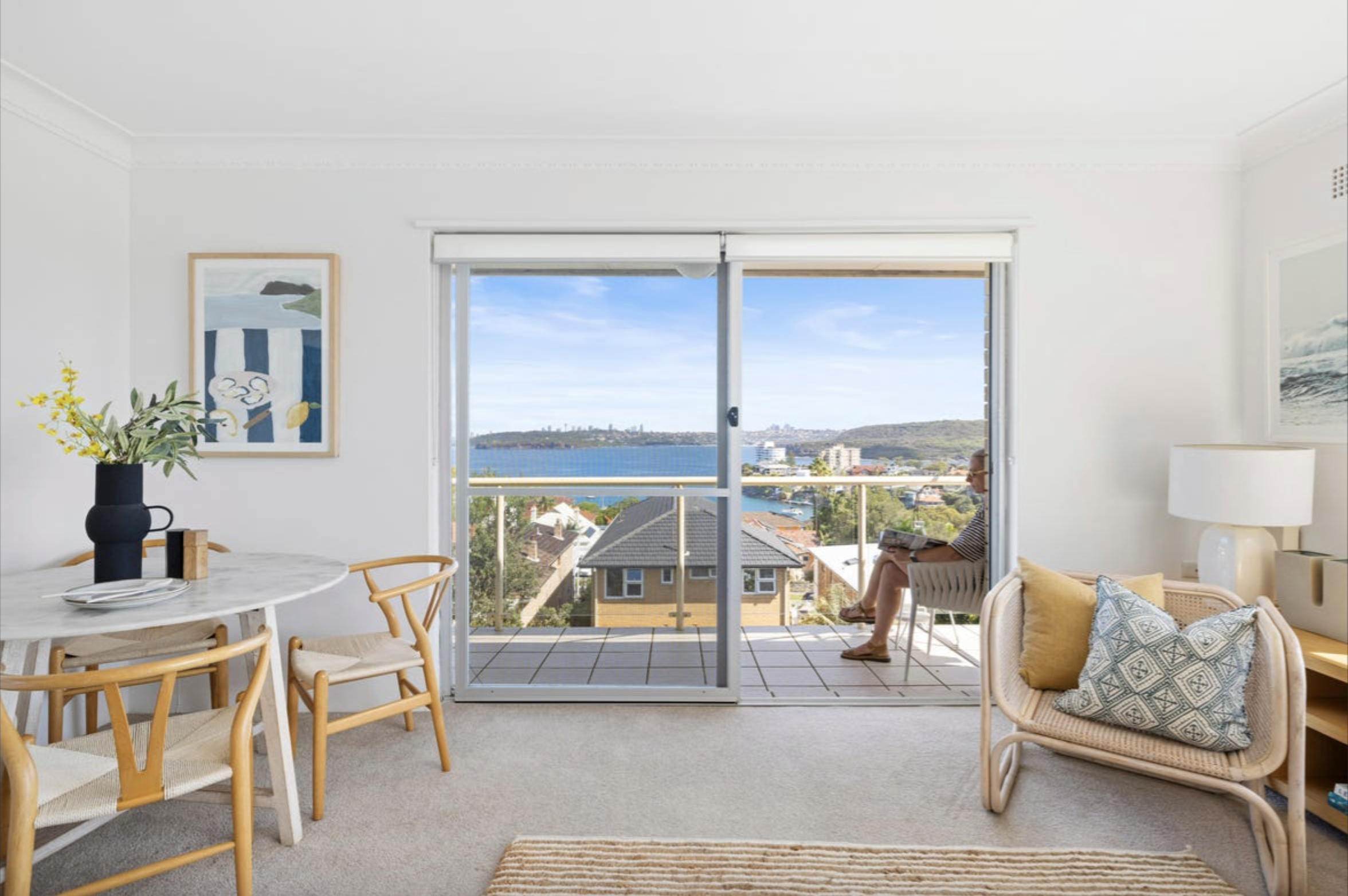 Bright living room with a marble dining table, wooden chairs, a vase of yellow flowers, and a sliding glass door opening to a coastal view.
