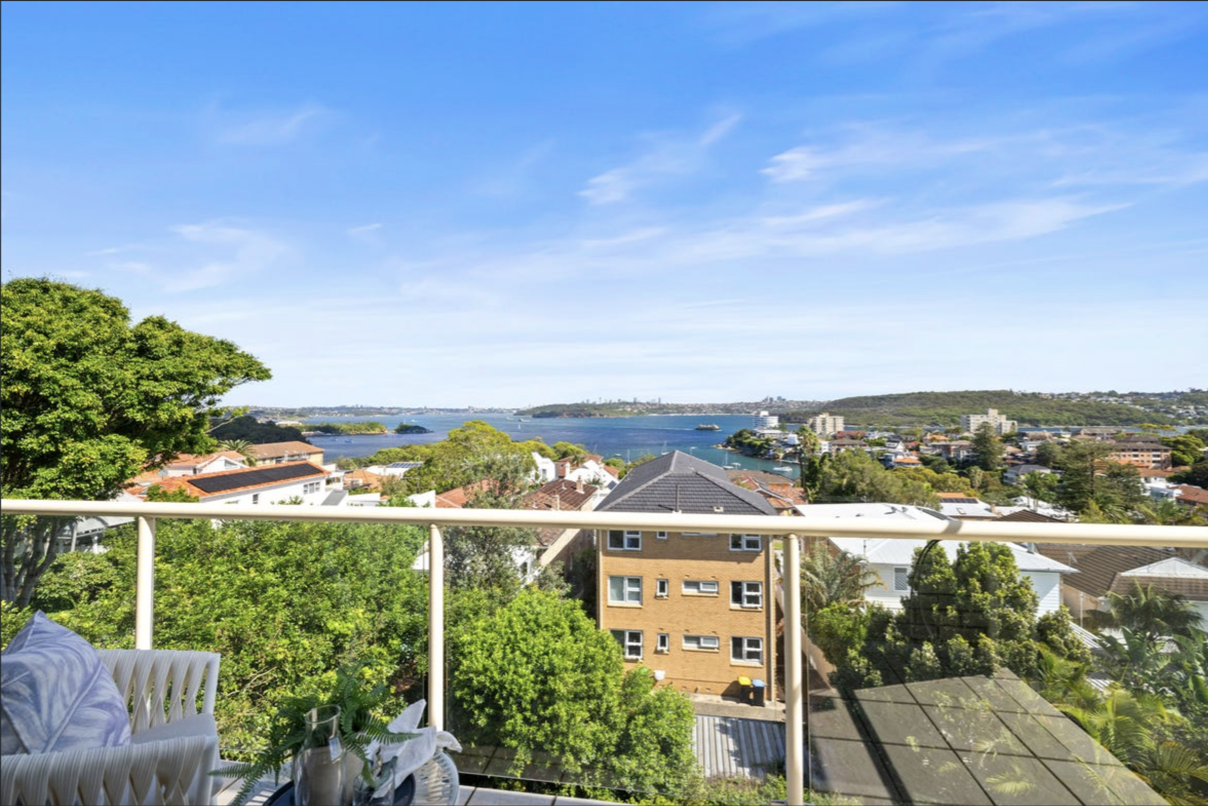 Balcony view over a coastal town with red-tiled roofs, green trees, and a blue bay under a clear sky.
