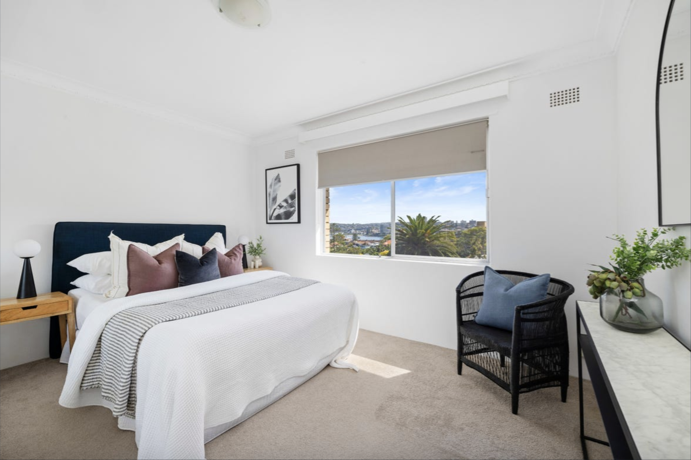 Bright, minimalist bedroom with a white palette, navy headboard, and striped bedding; window view of a sunny landscape with plants nearby