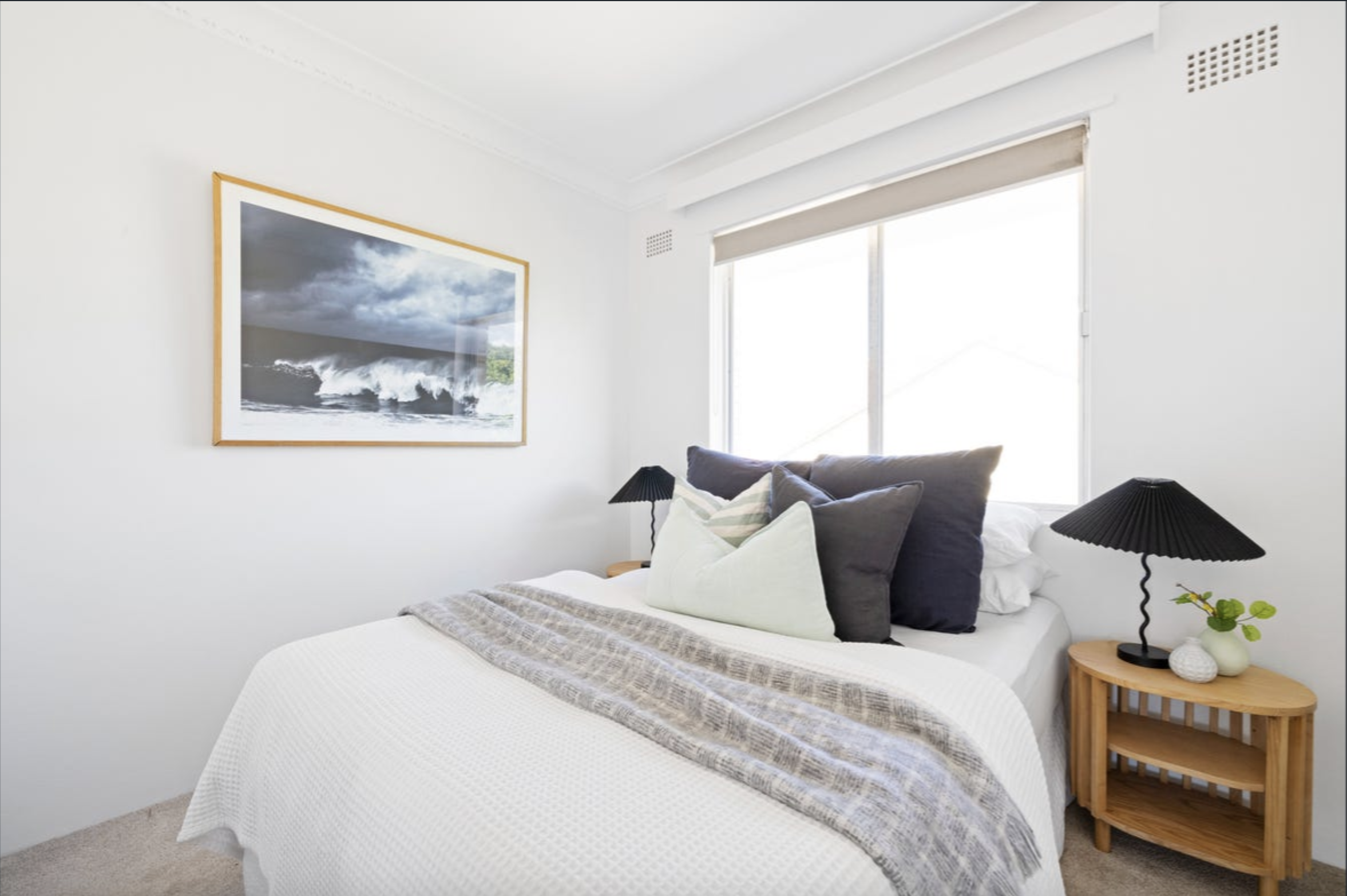 Bright bedroom with a white bed, multiple pillows, and a coastal framed artwork on the wall beside a large window.