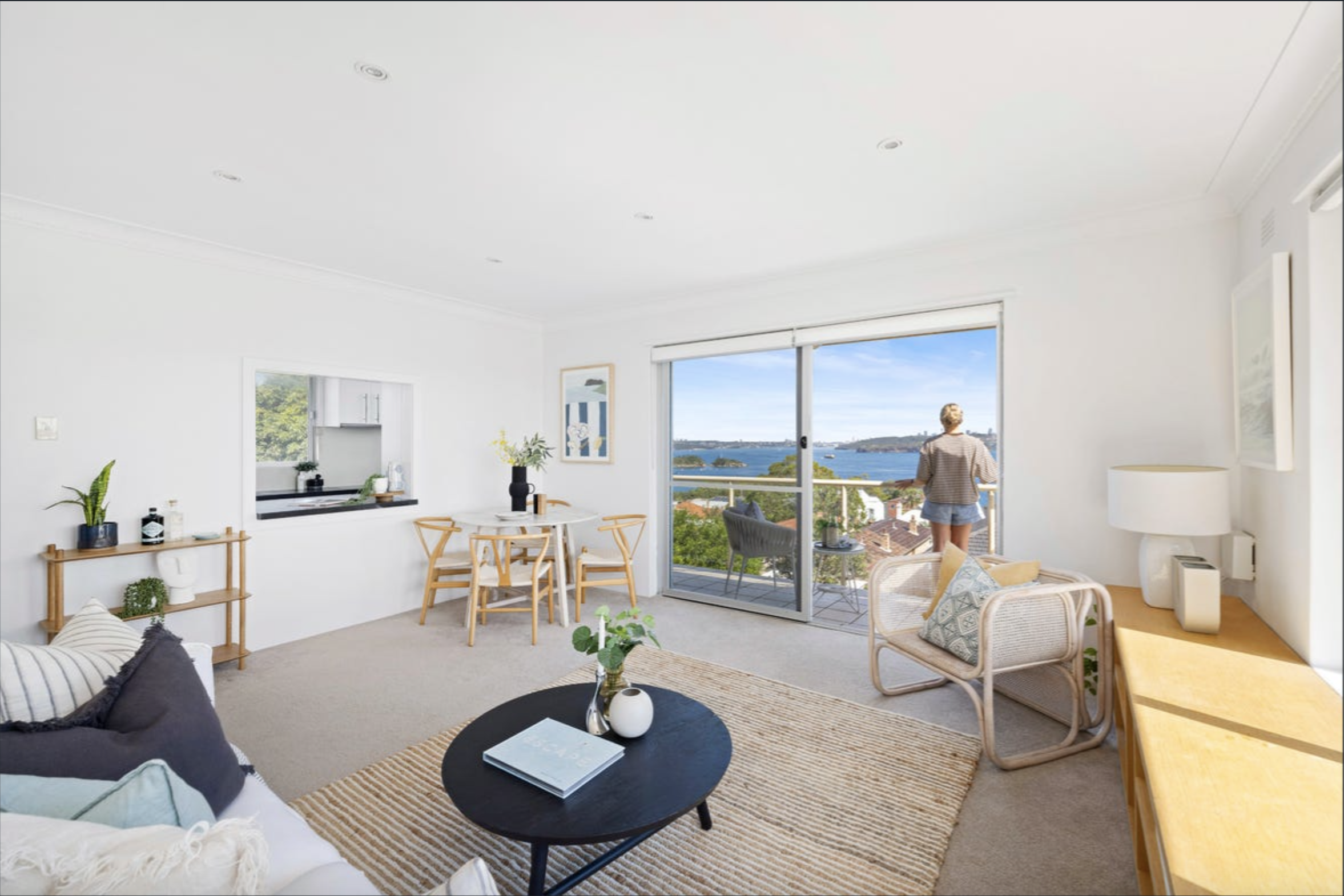 Bright living room with white walls, beige rug, circular black coffee table, and a person standing on the balcony overlooking the ocean.