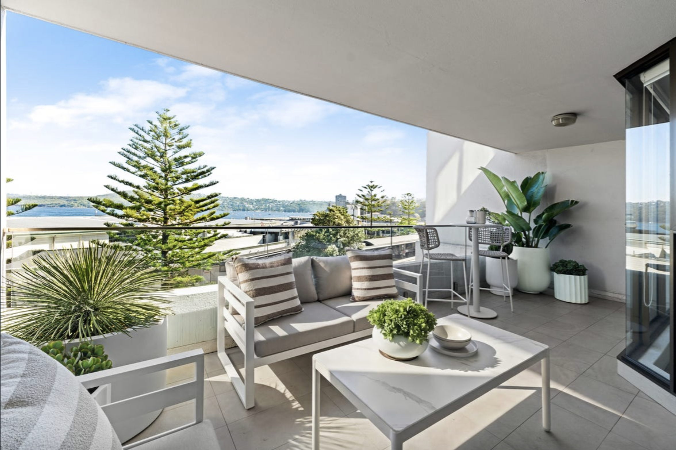 Modern balcony seating area with white furniture and striped cushions, potted plants, and a glass railing overlooking water and distant hills under a blue sky