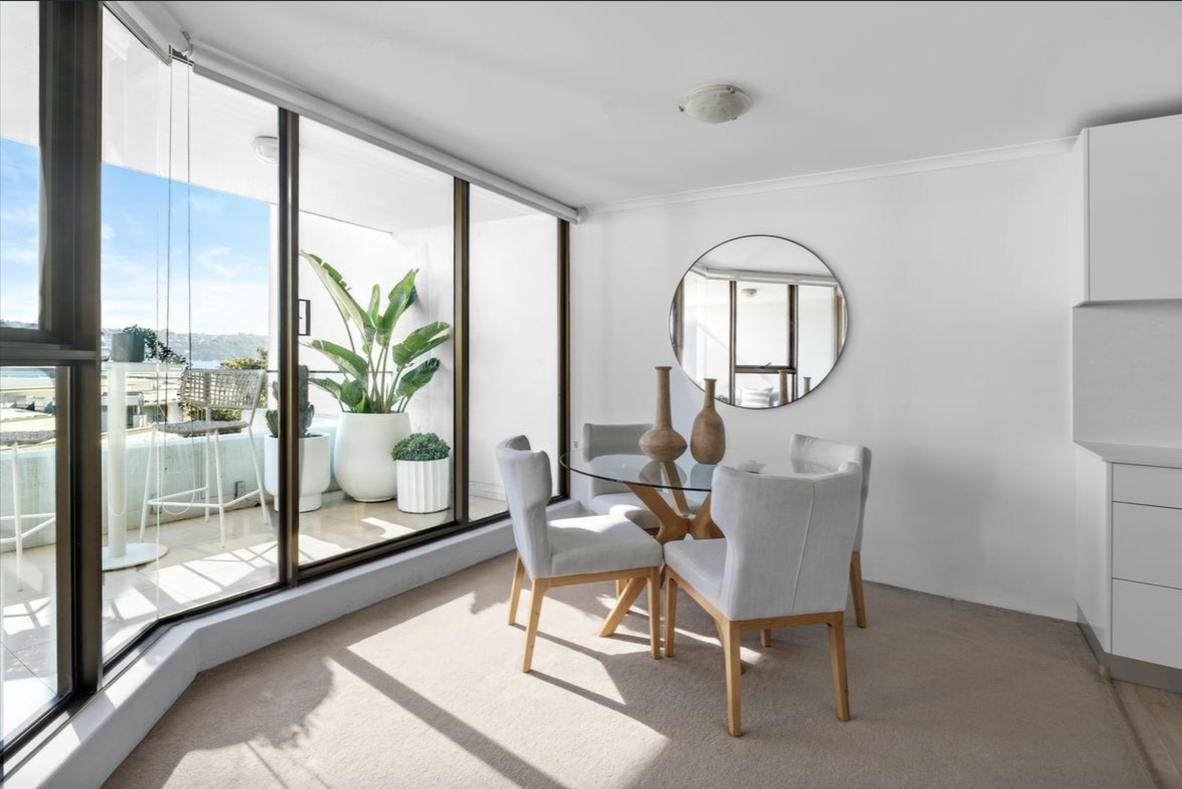 Bright modern dining area with a glass wall and balcony outside, white chairs around a round glass table, and large potted plants nearby.