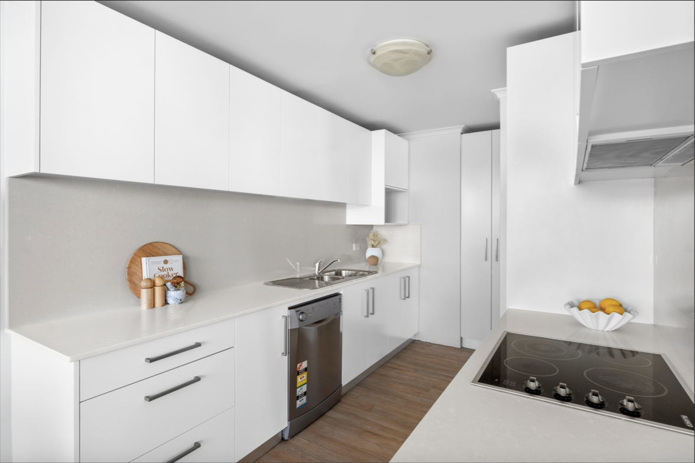 Bright white kitchen with glossy cabinets, a stainless steel dishwasher, and a black glass stovetop against a light-gray island countertop.