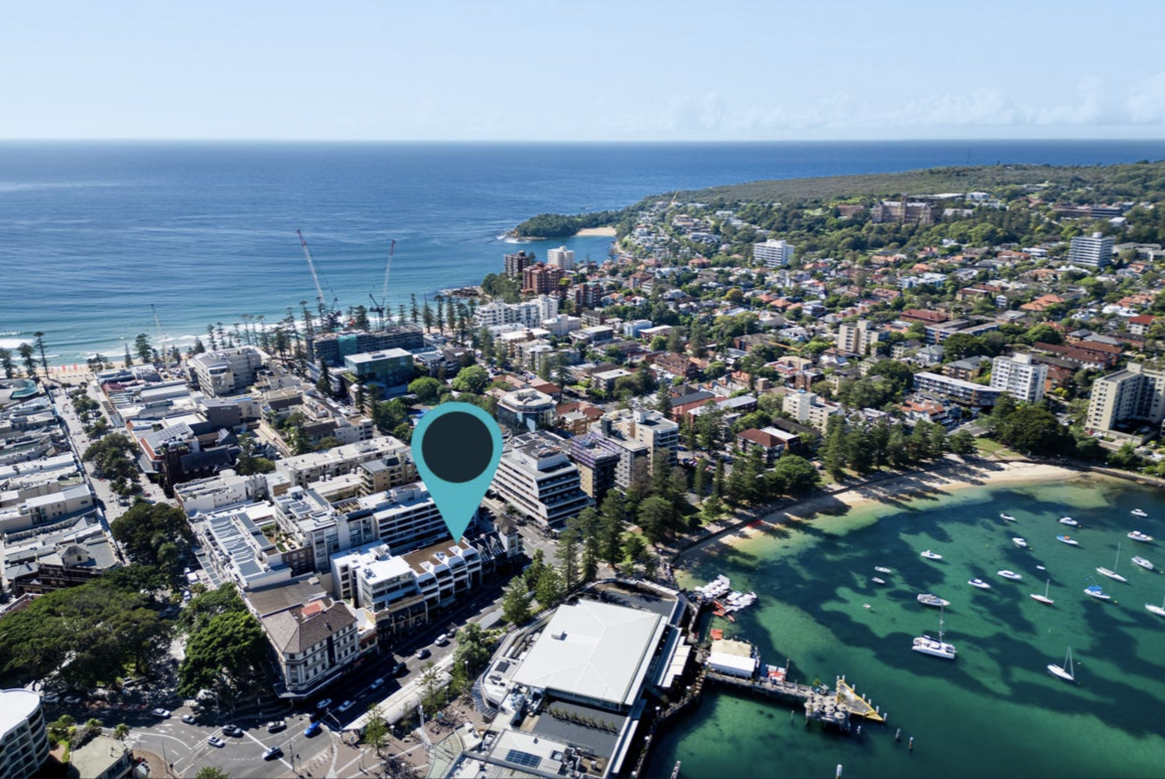 Aerial view of a coastal city with a marina, mid-rise buildings, and green hills in the distance; a teal map pin marks a location on the cityscape.