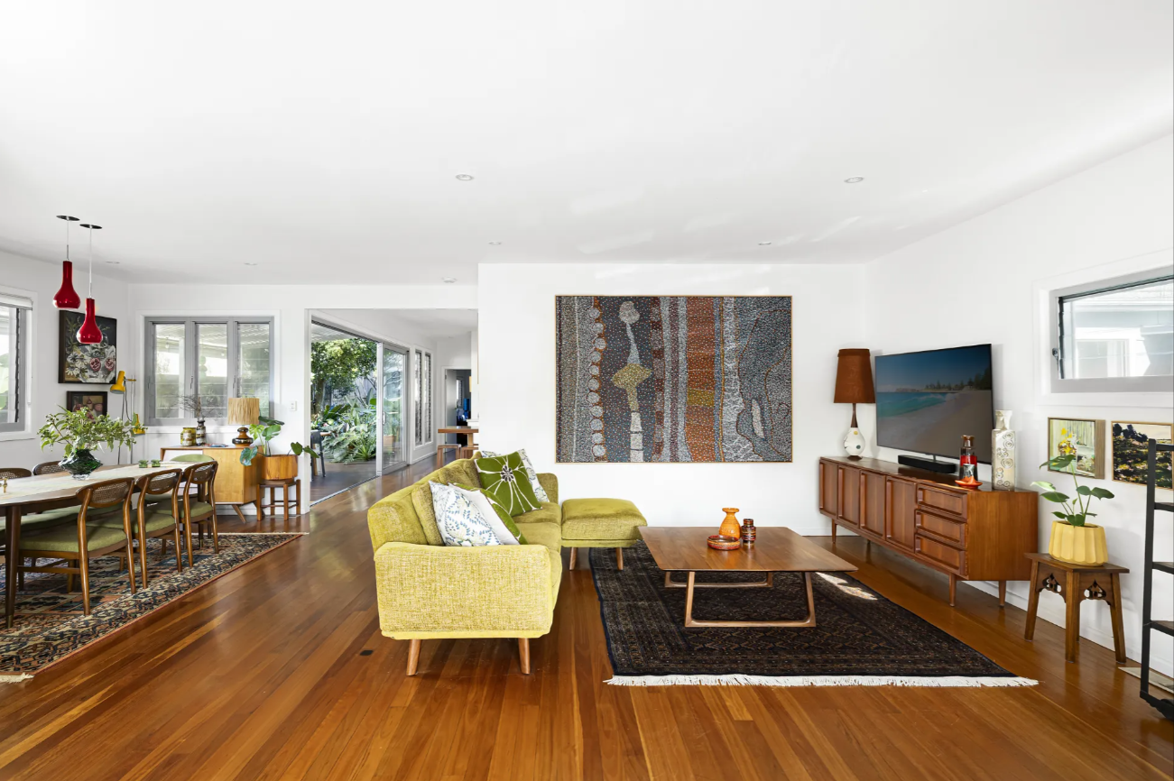 Open-concept living and dining room with a yellow sofa, wooden floors, and vintage furniture; large wall art above a rug-covered coffee table area.
