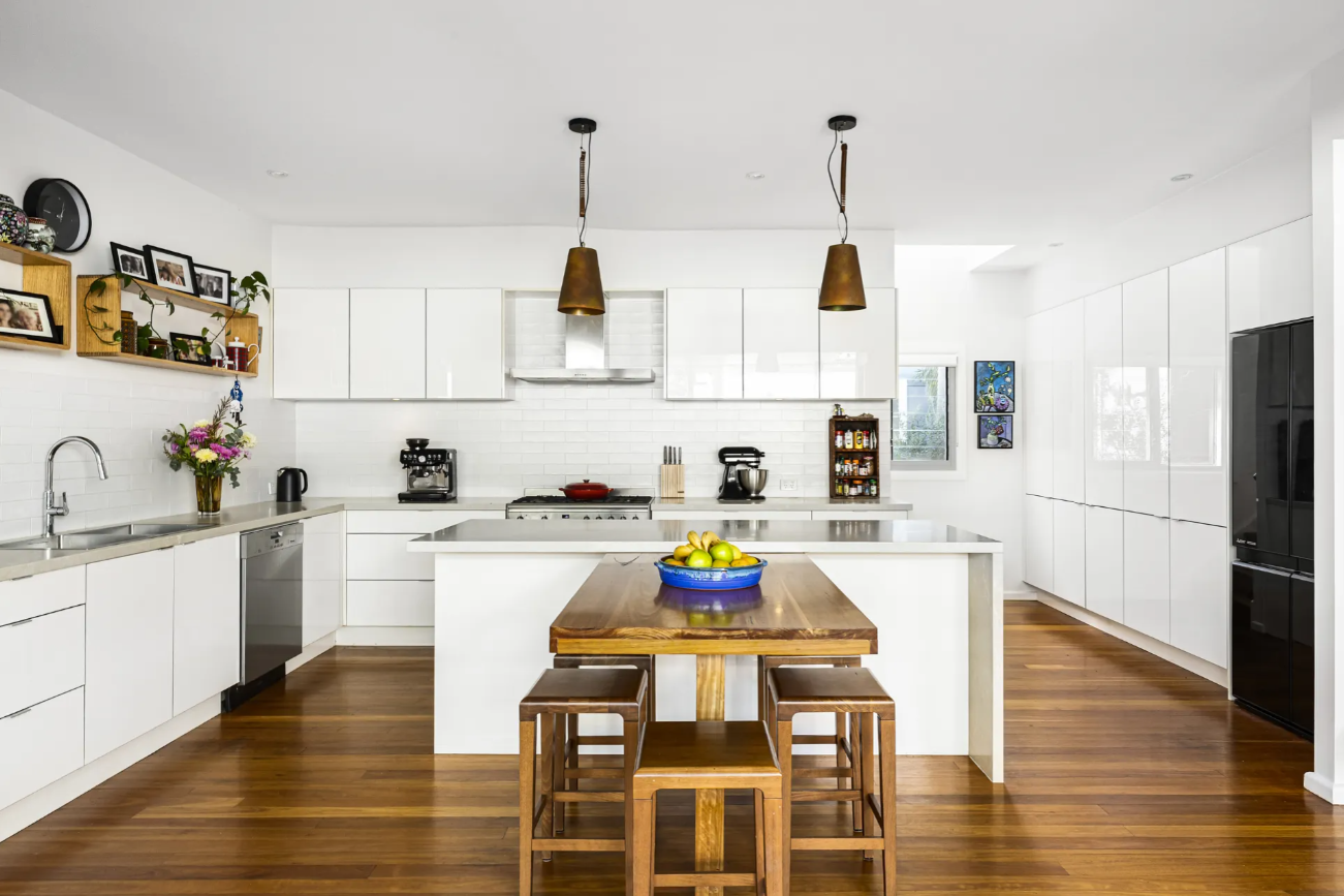 Modern white kitchen with an island, wooden dining table in the center, and a bowl of fruit on the table.