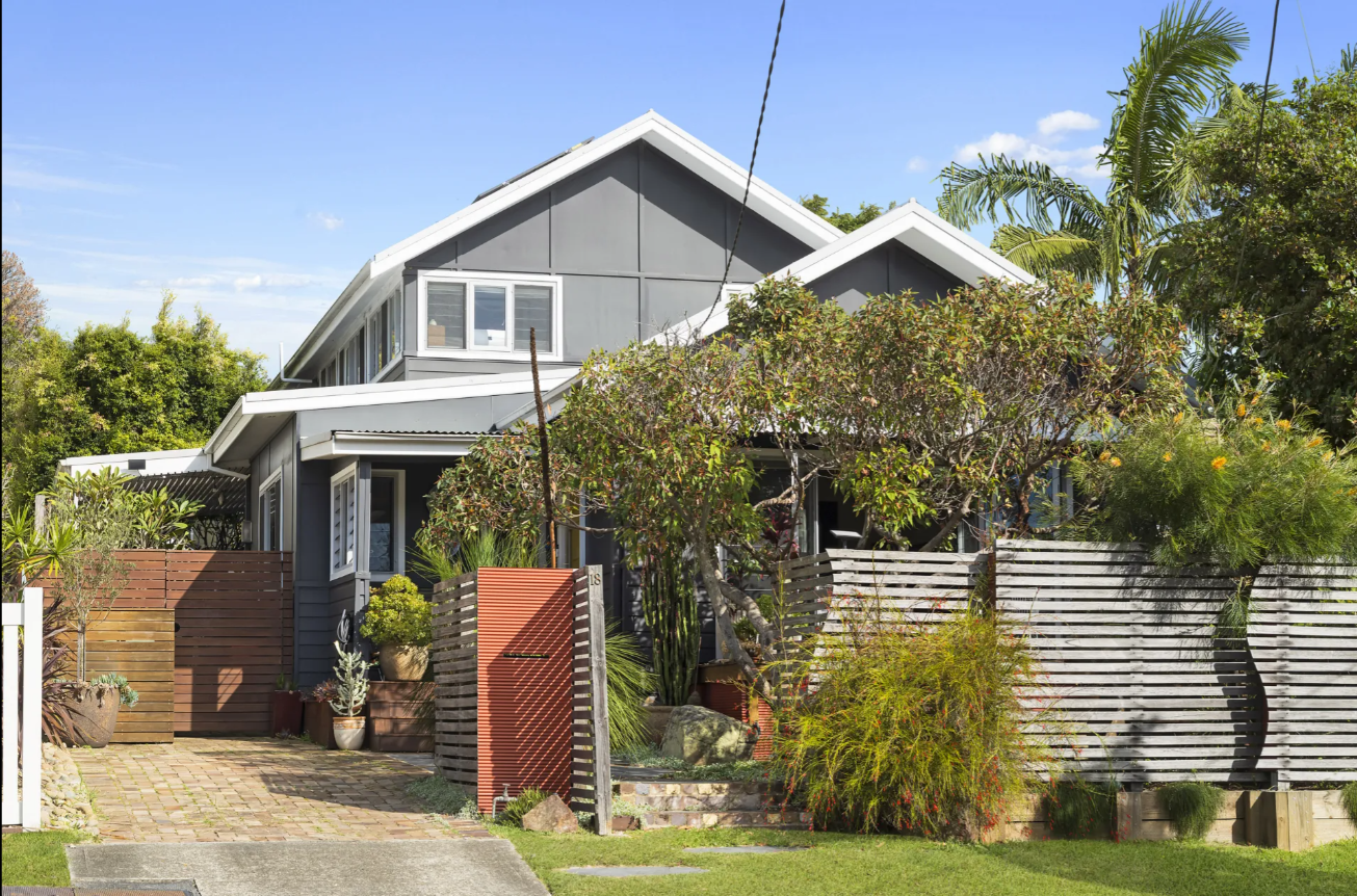Two-story gray house with white trim and gabled roofs, front yard with trees and a wooden slat fence and gate.