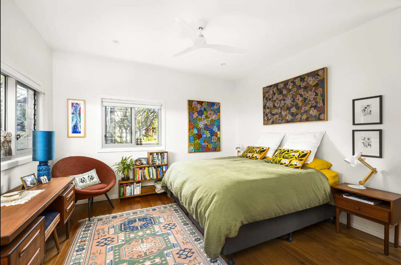 Bright bedroom with a green duvet bed, colorful wall art, and bedside tables on a wooden floor; a desk, chair, and bookshelf line the left wall under windows.