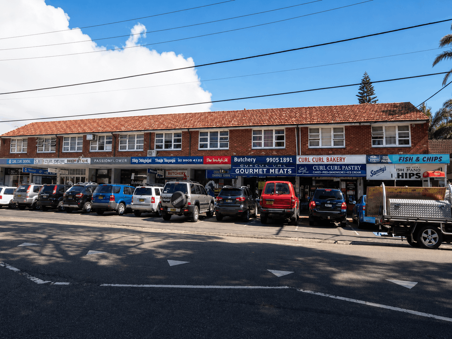 Curl Curl retail strip with shops like bakery, butchery, and fish and chips under brick building, cars outside.