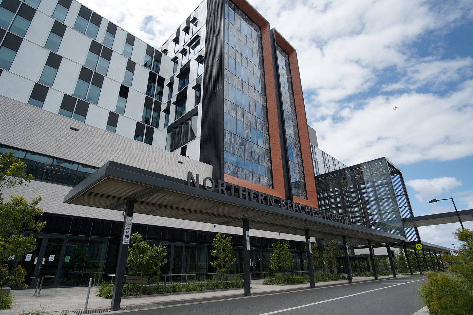 Northern Beaches Hospital, a modern glass building under a cloudy sky, with trees and covered walkway in front.