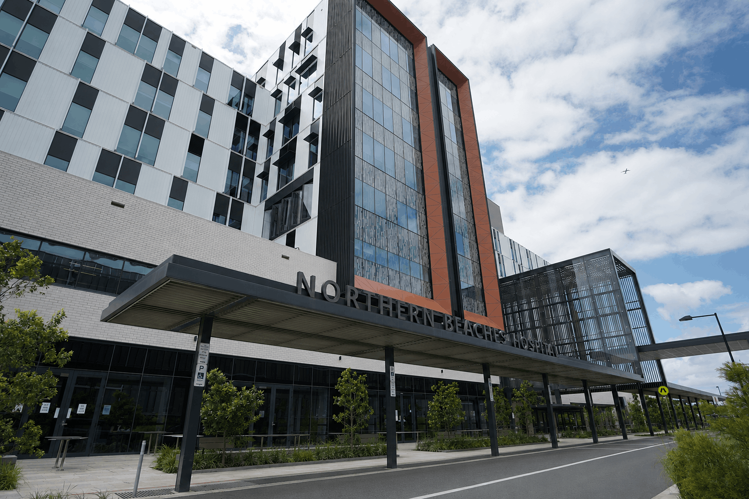 A modern multi-story hospital with large windows and covered entrance labeled “Northern Beaches Hospital” under cloudy sky.