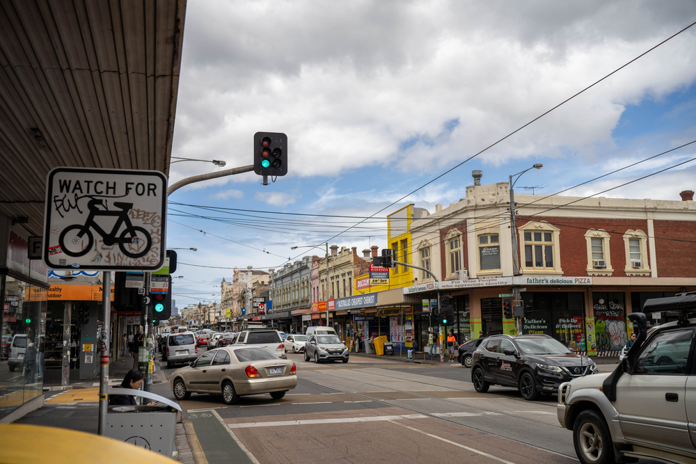 Busy urban street on Sydney Road with cars, shops, bicycle warning sign, green traffic lights, and pedestrians.