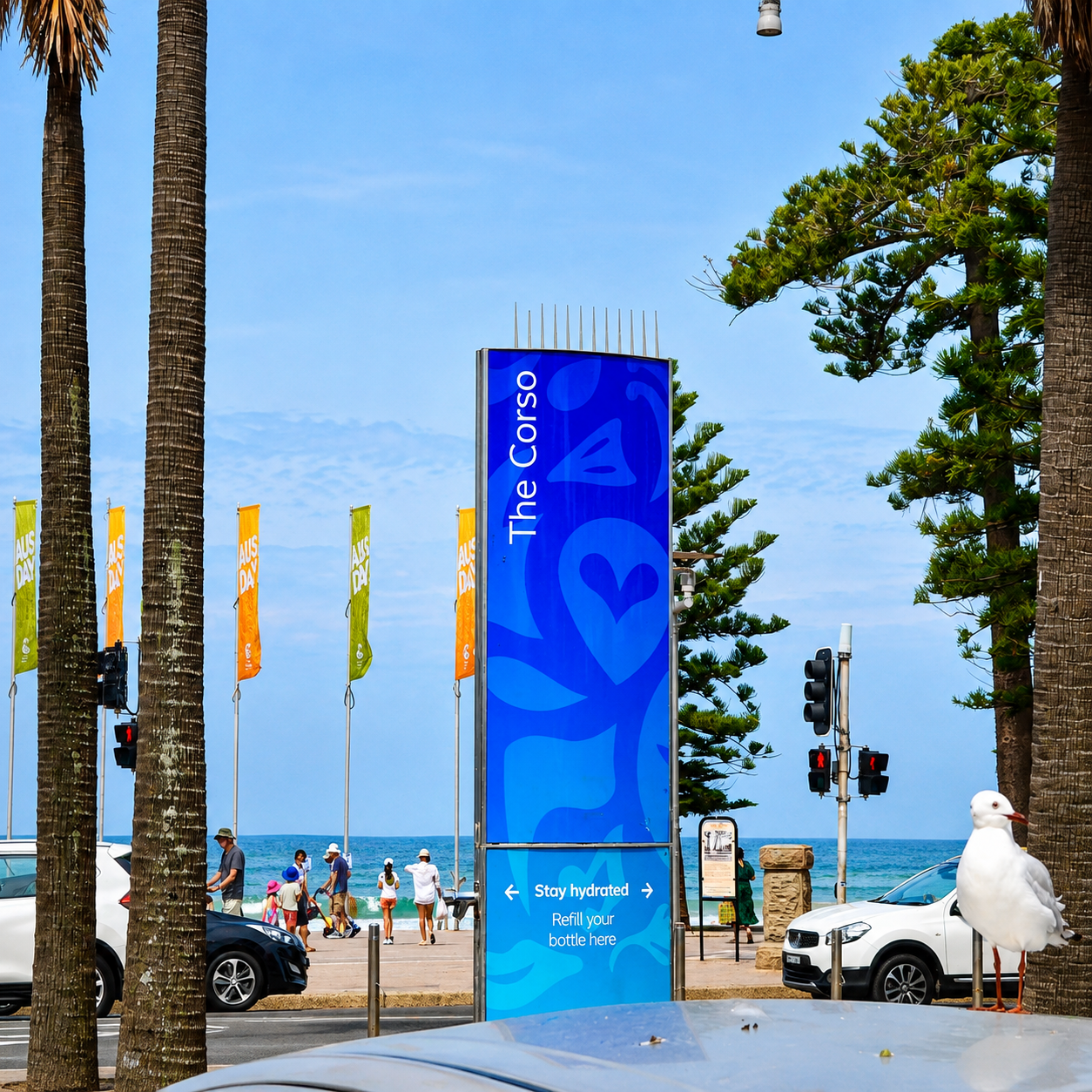 The Corso beachside sign with people, cars, a seagull, trees and ocean views in Manly.